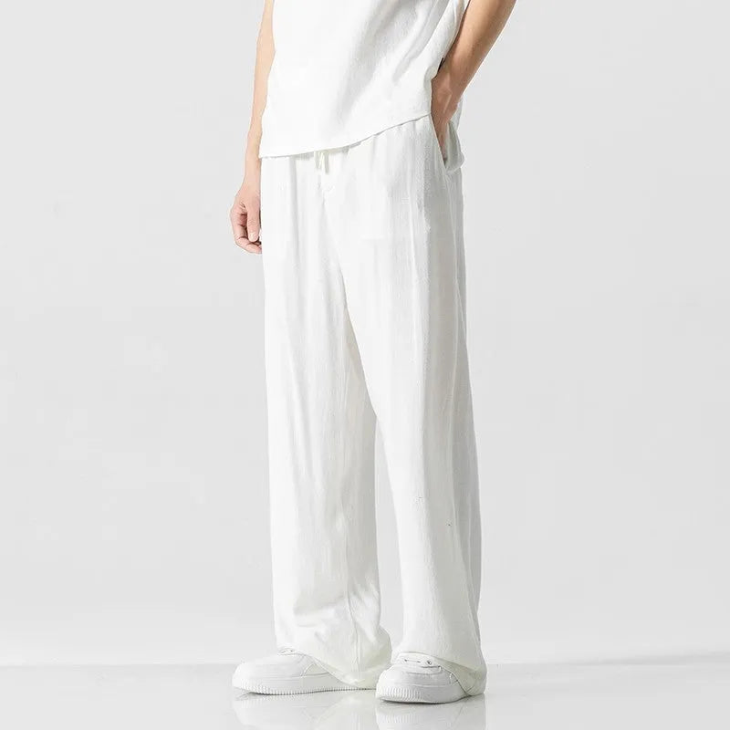 Men's beige linen-style trousers with a loose straight-leg cut and drawstring waist, shown in a casual setting.