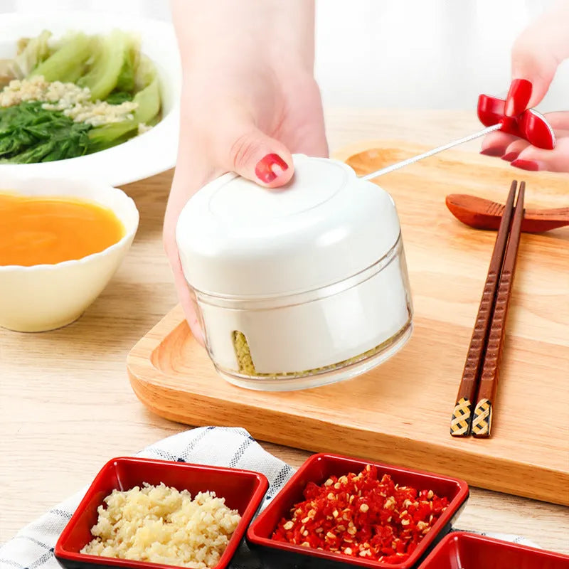 White manual food chopper with stainless steel blades on a magnet, shown with garlic and vegetables.