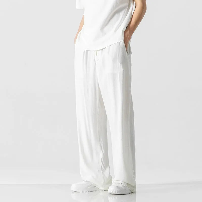 Men's beige linen-style trousers with a loose straight-leg cut and drawstring waist, shown in a casual setting.