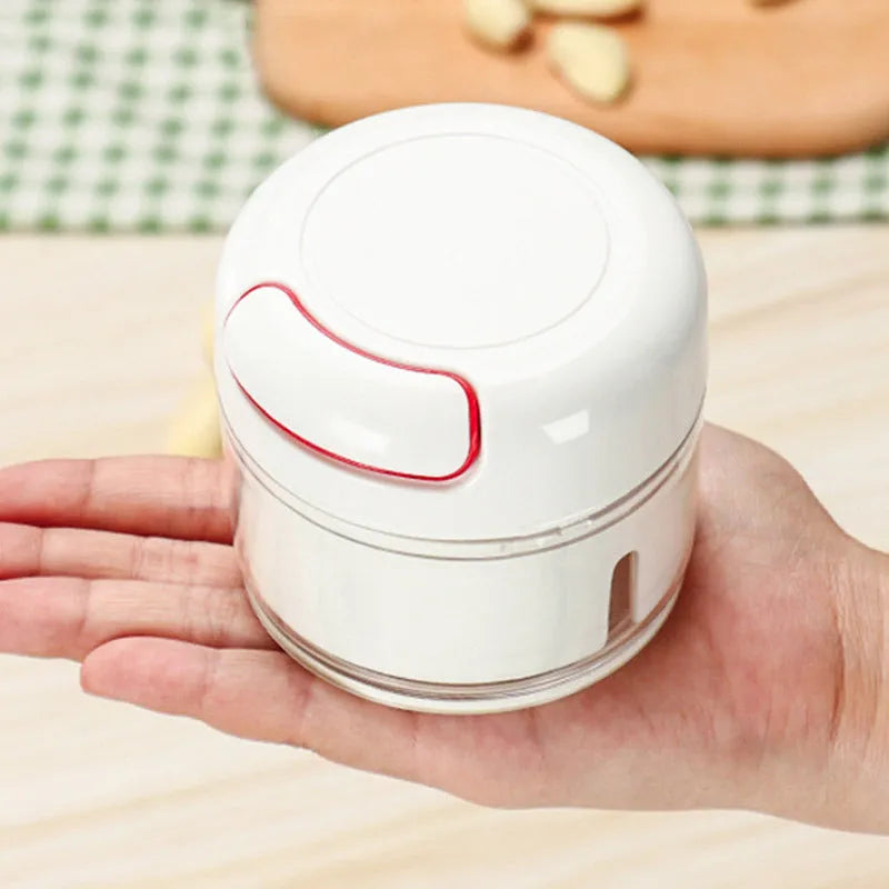 White manual food chopper with stainless steel blades on a magnet, shown with garlic and vegetables.