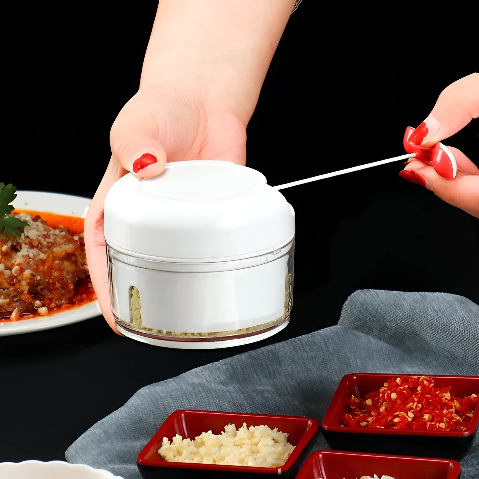White manual food chopper with stainless steel blades on a magnet, shown with garlic and vegetables.