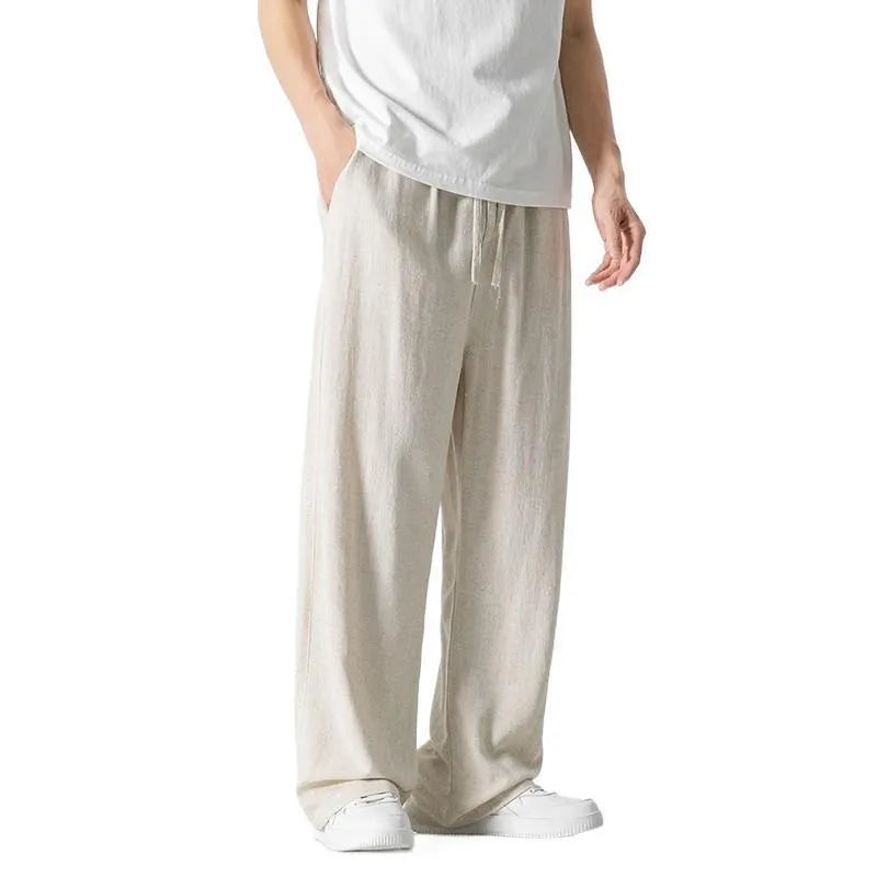 Men's beige linen-style trousers with a loose straight-leg cut and drawstring waist, shown in a casual setting.