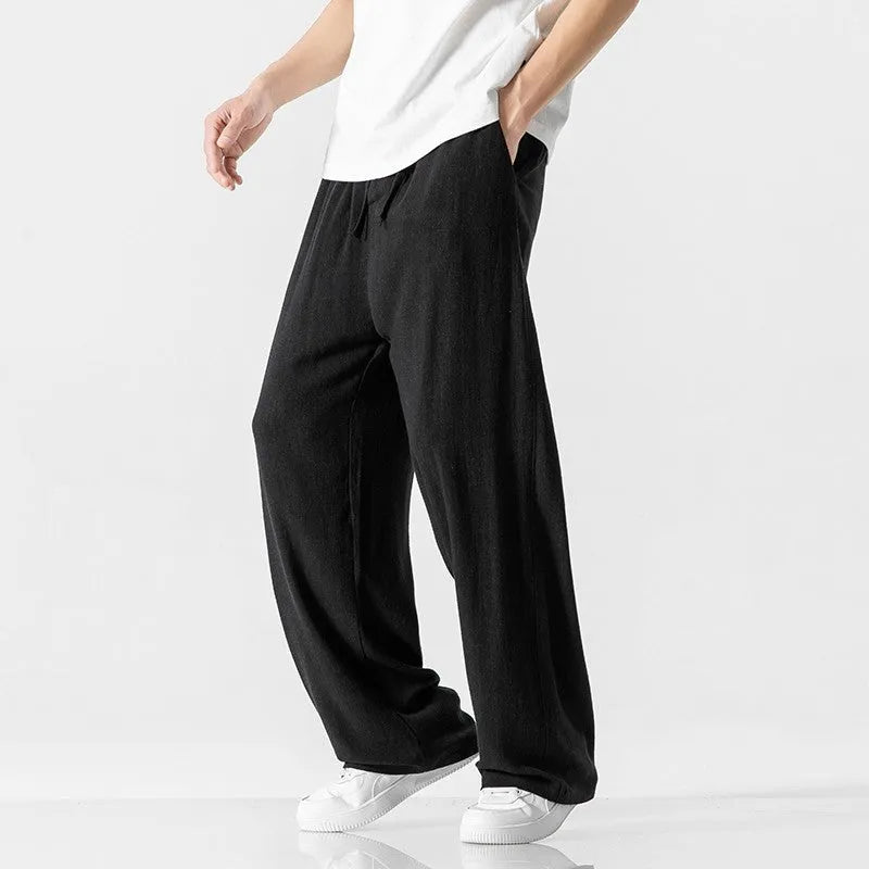 Men's beige linen-style trousers with a loose straight-leg cut and drawstring waist, shown in a casual setting.