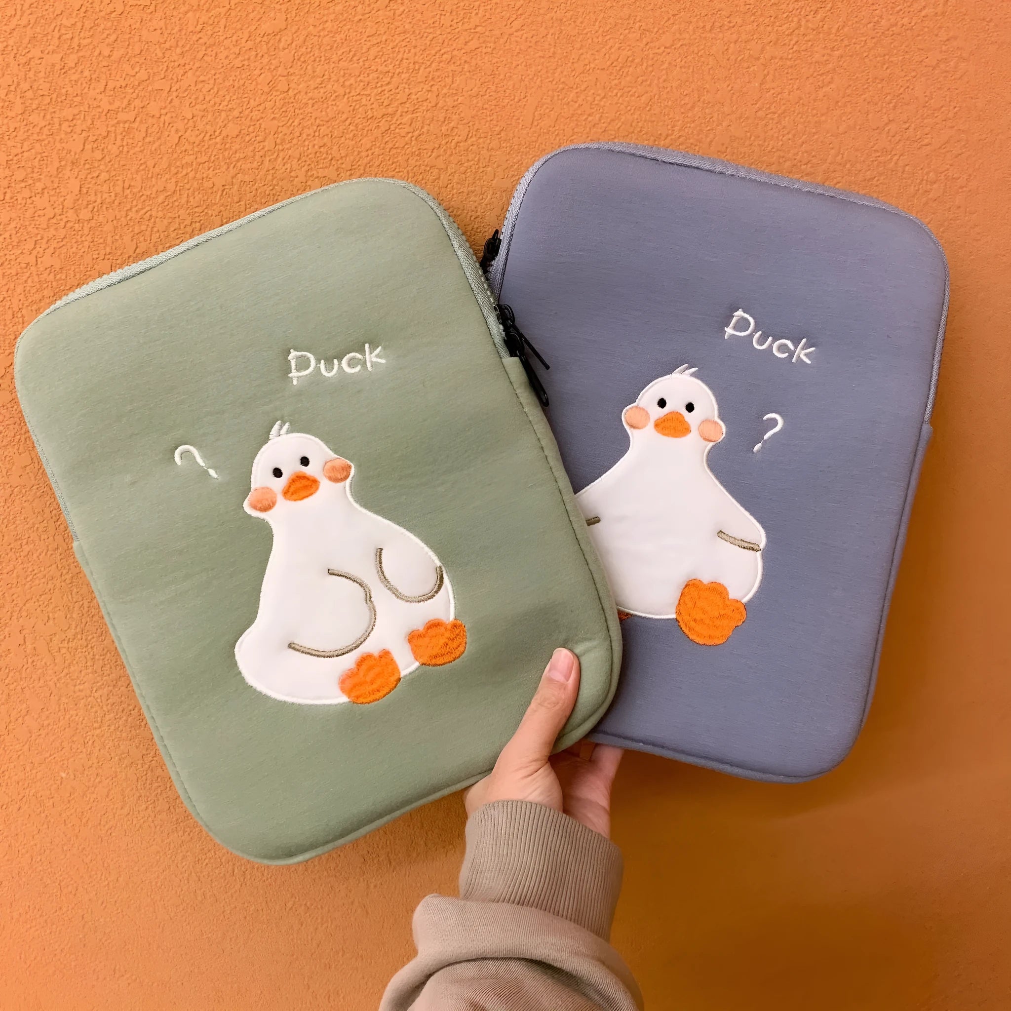 Blue cotton tablet case with embroidered cartoon duck design and a zipper, shown next to a green version.