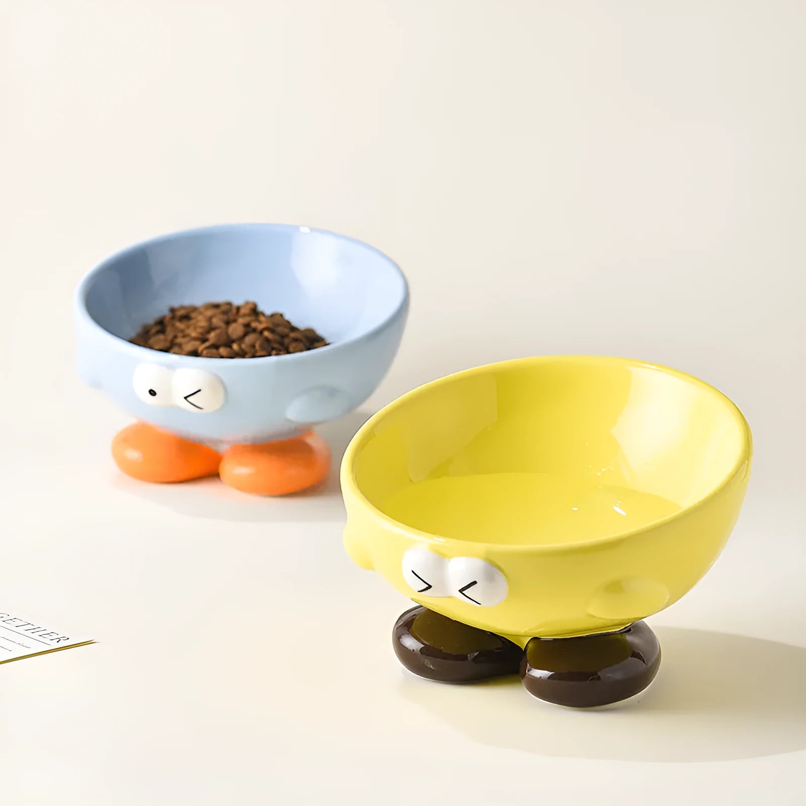 Blue and yellow ceramic slanted pet food bowls with animal face expressions for cervical spine support.