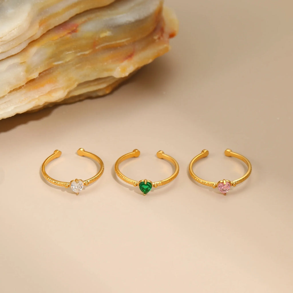 Open-ended geometric stainless steel ring with a heart-shaped green zirconium inlay on a grey background.