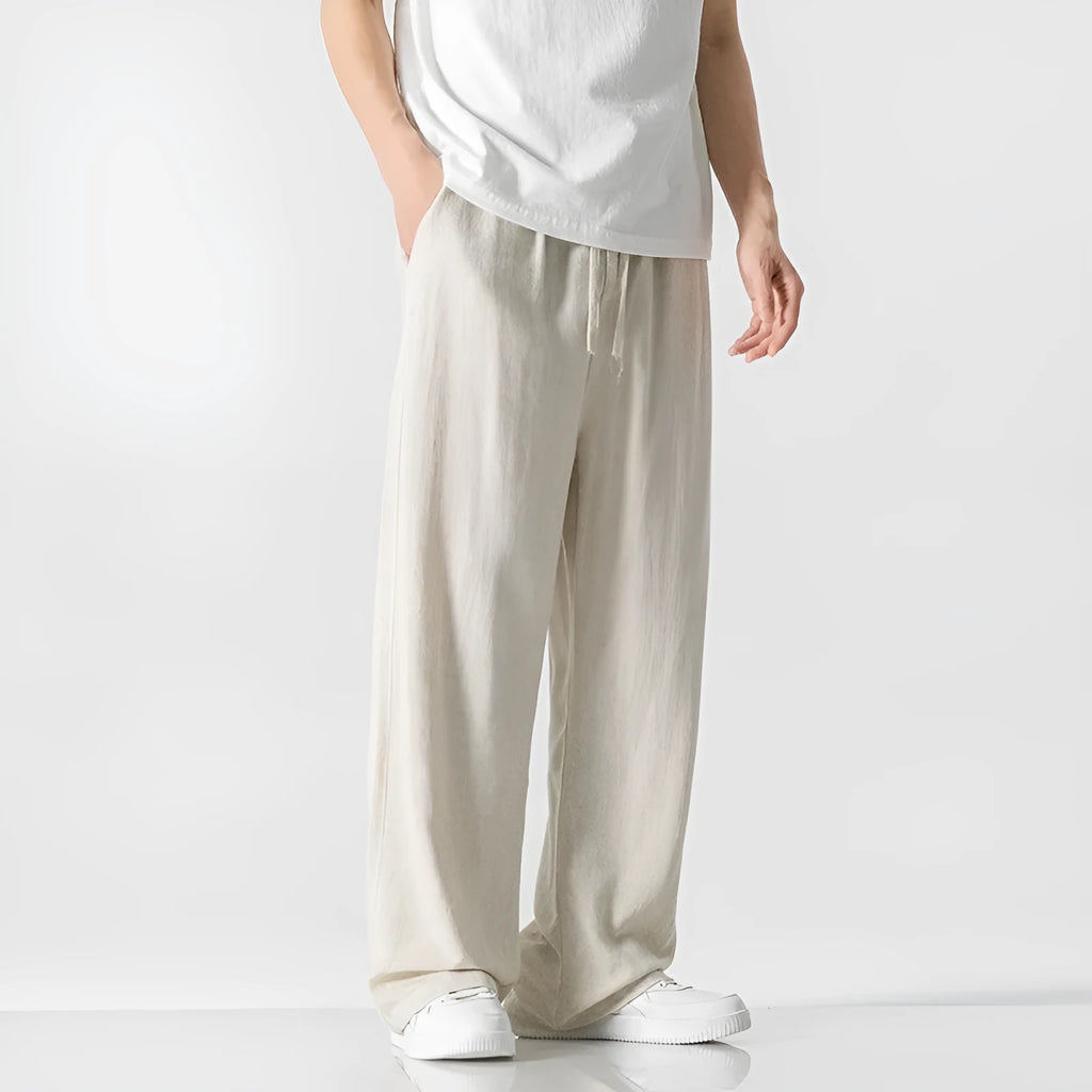 Men's beige linen-style trousers with a loose straight-leg cut and drawstring waist, shown in a casual setting.