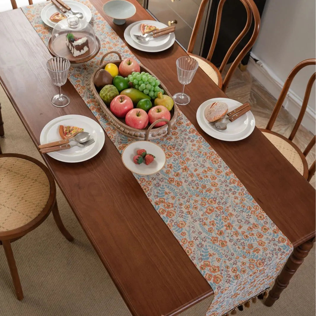 Beige jacquard table runner with floral pattern and long tassels on a wooden table.