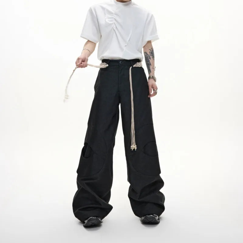Men's dark coloured loose-fit straight-leg cotton trousers with decorative tassel and pocket zipper details.