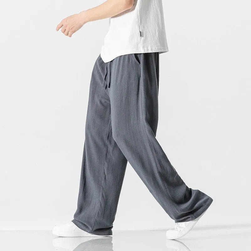 Men's beige linen-style trousers with a loose straight-leg cut and drawstring waist, shown in a casual setting.